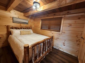 Un dormitorio de cabaña de madera con cama, cabecera, ventana con persianas, lámpara de techo e imagen enmarcada en la pared.