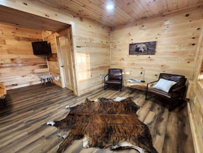 Interior de uma cabana de madeira com um tapete de pele grande, duas poltronas de couro, uma televisão e um quadro de urso na parede.