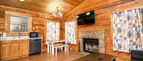 A wooden cabin interior with a kitchen, dining area, fireplace, and a TV mounted on the wall.