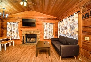 Interior de una cabaña de troncos con chimenea, un sofá, un televisor y paredes de madera.