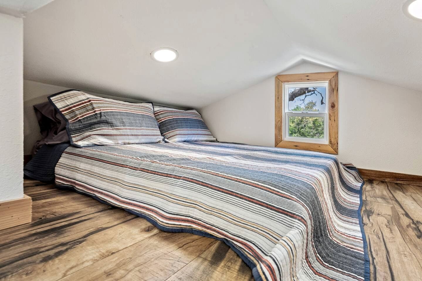 A loft-style bedroom with a sloped ceiling, a large bed with striped bedding, a wooden headboard, a framed picture on the wall, and a window with a view of greenery.