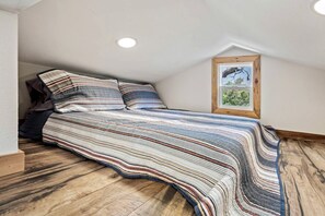 Un dormitorio estilo loft con techo inclinado, una cama grande con ropa de cama a rayas, un cabecero de madera, un cuadro enmarcado en la pared y una ventana con vistas a la vegetación.
