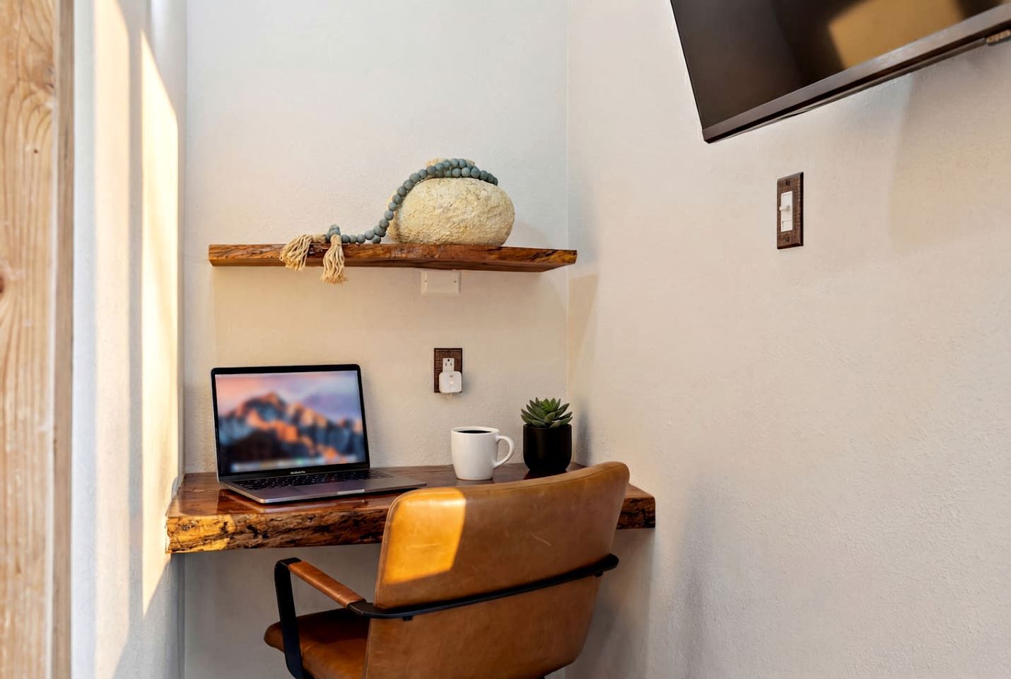 A workspace with a laptop, a chair, a shelf with a plant, and a wall-mounted TV.