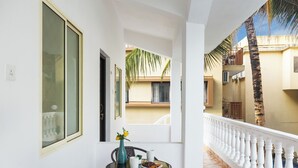 Un balcon doté d’une balustrade blanche, une table ronde avec un vase et une assiette de nourriture, et une vue sur les bâtiments voisins et un palmier.