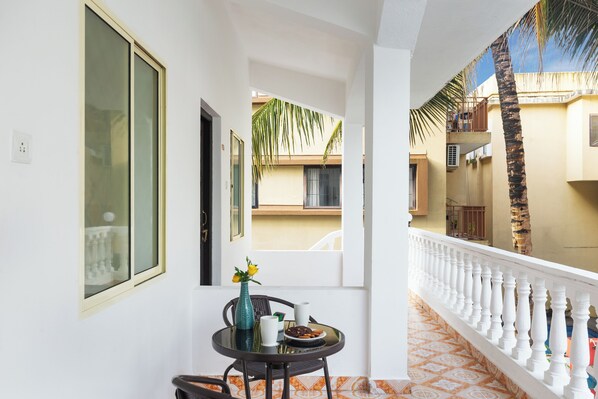 A balcony with a white balustrade, a round table with a vase and a plate of food, and a view of neighboring buildings and a palm tree. - 8fold by LaRiSa Siolim (Siolim)