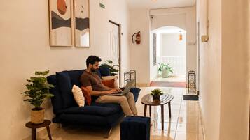 Um homem sentado em um sofá azul escuro em um corredor bem iluminado, com um laptop, uma pequena mesa redonda com uma planta e um vaso com planta ao lado do sofá.