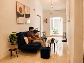 Um homem sentado em um sofá azul escuro em um corredor bem iluminado, com um laptop, uma pequena mesa redonda com uma planta e um vaso com planta ao lado do sofá.