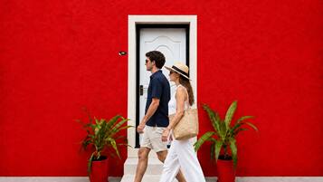 A couple walking past a red wall with a white door, two potted plants, and a small sign with the number 106.