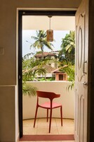 Puerta abierta que da a un balcón con una silla roja, una lámpara colgante y vistas a palmeras y edificios.