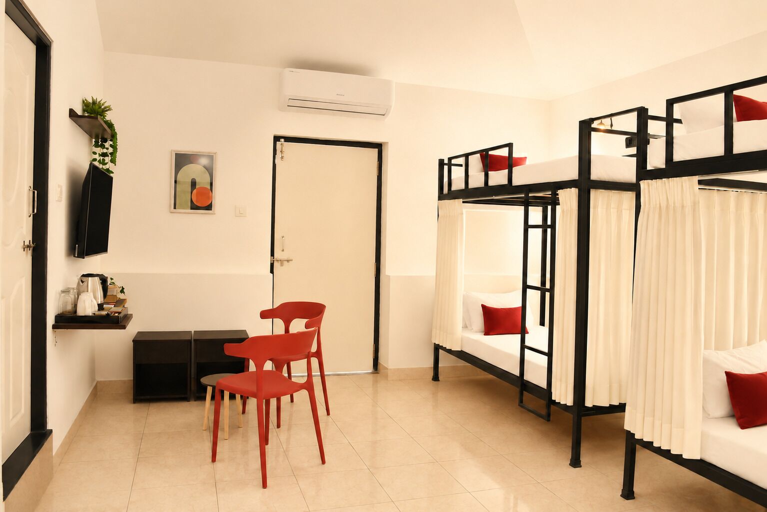 A room with bunk beds, a desk with chairs, a door, and a wall-mounted shelf with items.