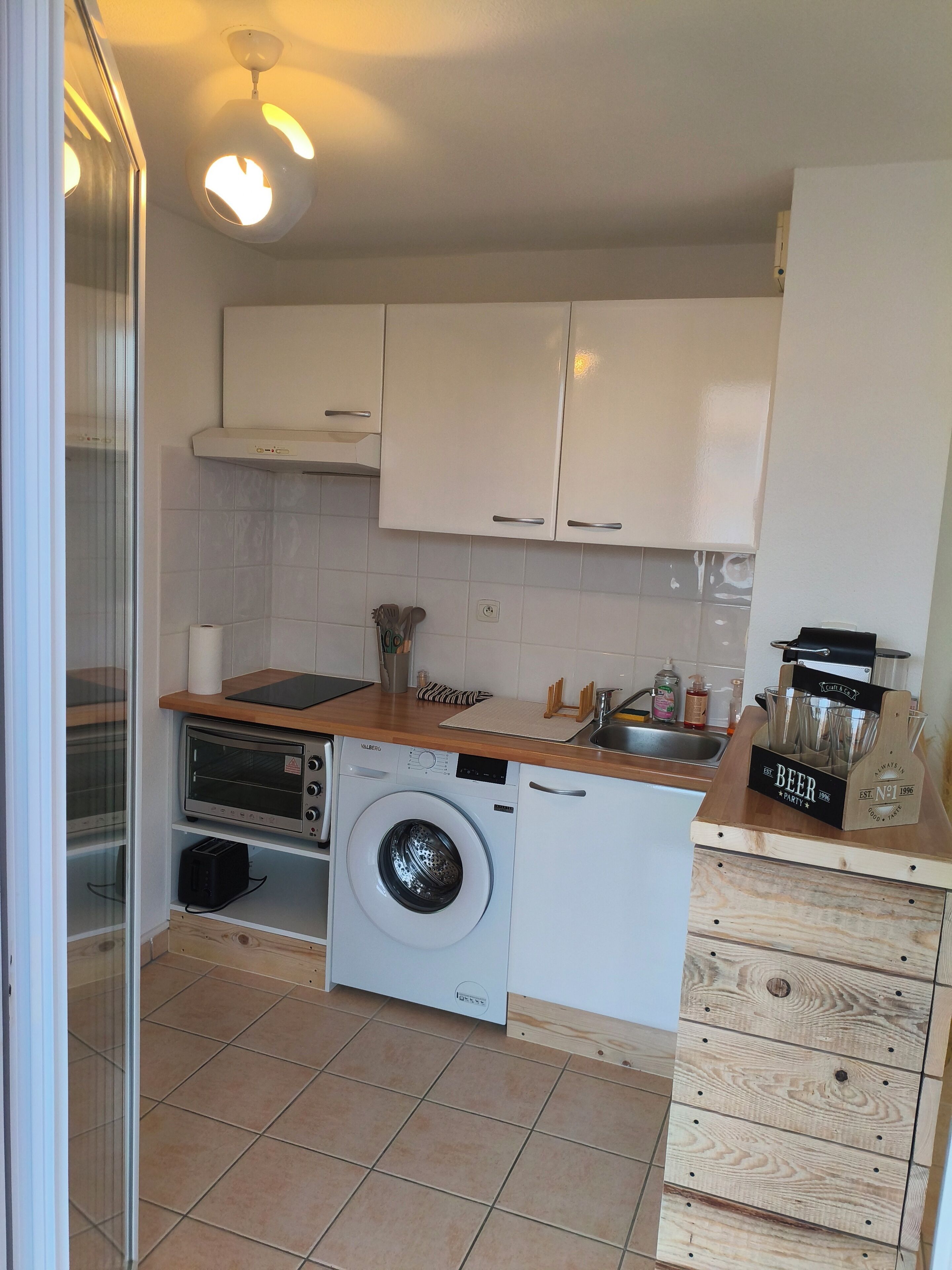 Cocina compacta con muebles blancos, lavadora, tostadora y encimera de madera con grifo de cerveza.