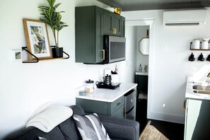 Une cuisine compacte équipée d’un four à micro-ondes, d’une machine à café et d’un évier. Un petit coin salon avec un canapé et une plante en pot est également disponible.