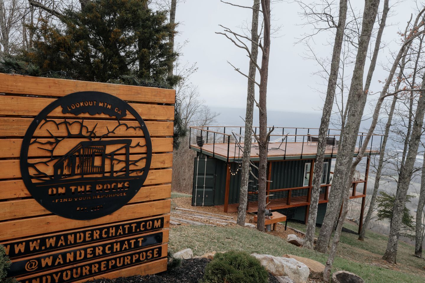 Un panneau en bois avec un logo et du texte, un chalet doté d’une terrasse, et une zone boisée offrant une vue sur l’eau.