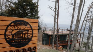 Ein Holzschild mit Logo und Text, eine Blockhütte mit Veranda und ein bewaldetes Gebiet mit Blick auf das Wasser.