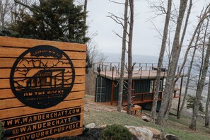 A wooden sign with a logo and text, a cabin with a deck, and a forested area with a view of the water.