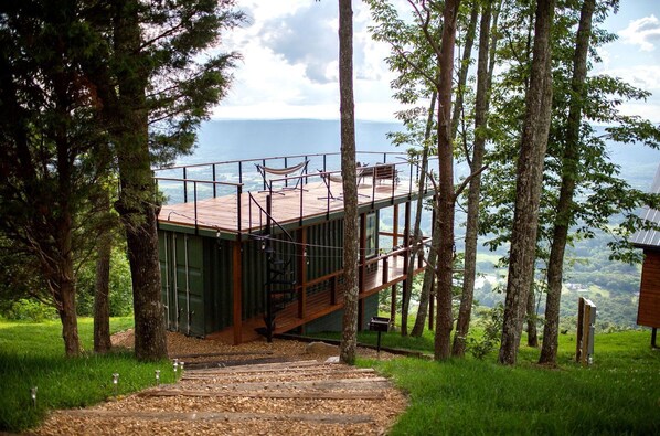 A treehouse with a wooden deck and stairs, surrounded by trees and a view of the landscape. - Incredible Container with Rooftop and Beautiful Sunset Views in Georgia (Rising Fawn)