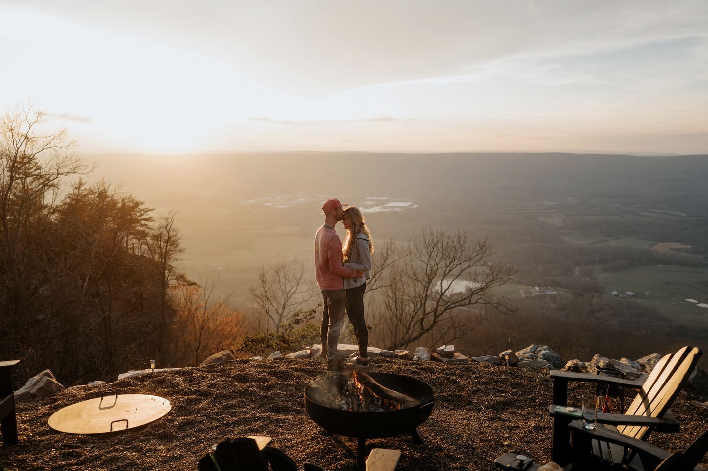 A couple standing by a campfire with a scenic view in the background.
