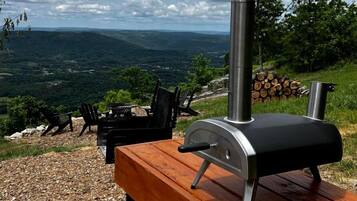 A barbecue grill with a chimney, a wooden table, and chairs on a gravel surface with a scenic view in the background.