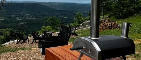 A barbecue grill with a chimney, a wooden table, and chairs on a gravel surface with a scenic view in the background.