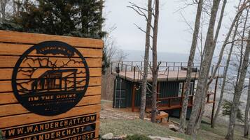 Ein Holzschild mit Logo und Text, eine Blockhütte mit Veranda und ein bewaldetes Gebiet mit Blick auf das Wasser.