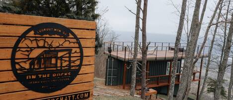 A wooden sign with a logo and text, a cabin with a deck, and a forested area with a view of the water.