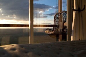 A wicker chair with a cushioned seat and back, positioned near a window with a view of a cloudy sky and a distant landscape.