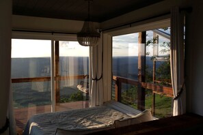 Une chambre à coucher avec un grand lit, un lustre et une vue sur un paysage, visible à travers de grandes fenêtres.
