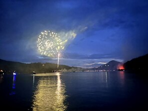 Ein Nachtblick auf einen See mit Feuerwerk am Himmel und Spiegelungen auf der Wasseroberfläche.