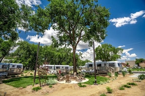 Un camping con vehículos recreativos, zona de fogata y árboles.