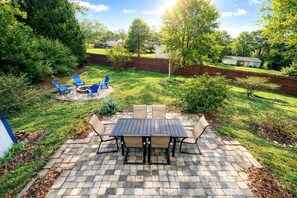 A patio with a table and chairs, surrounded by a brick border, with a grassy area and trees in the background.