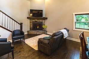 A living room with a stone fireplace, a television mounted above it, a staircase on the left, and a window on the right. - Sweet Private Cabin with Jacuzzi and Fire Pit in Ohio (Sugarcreek)
