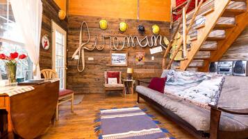 A cozy cabin interior with a wooden wall, a sofa, a chair, a small table, and a staircase.