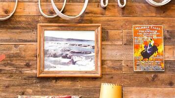 A rustic room with a wooden wall, a framed picture, a chair with a cushion, a small table with a lamp, and a bed.