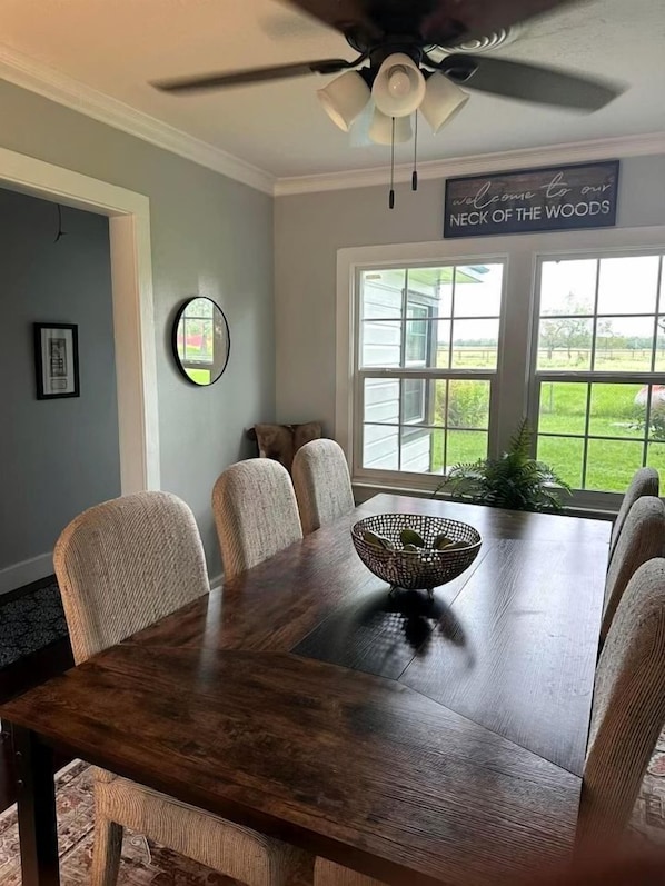 A dining room with a dark wooden table, six chairs, a ceiling fan, and a window with a view of greenery.