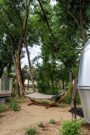 Una hamaca bajo un árbol, una parrilla y un baño químico en un área boscosa.