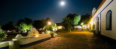 A cobblestone courtyard with a fountain, illuminated by street lamps and a full moon.