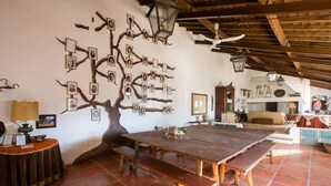 Un coin repas rustique avec une grande table et des bancs en bois, une fresque murale représentant un arbre agrémenté de photos, et un ventilateur de plafond.