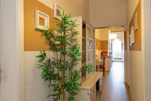 Un pasillo con una planta en maceta, cuadros en las paredes, un comedor con mesa y sillas, y una zona de cocina con armarios y encimera.