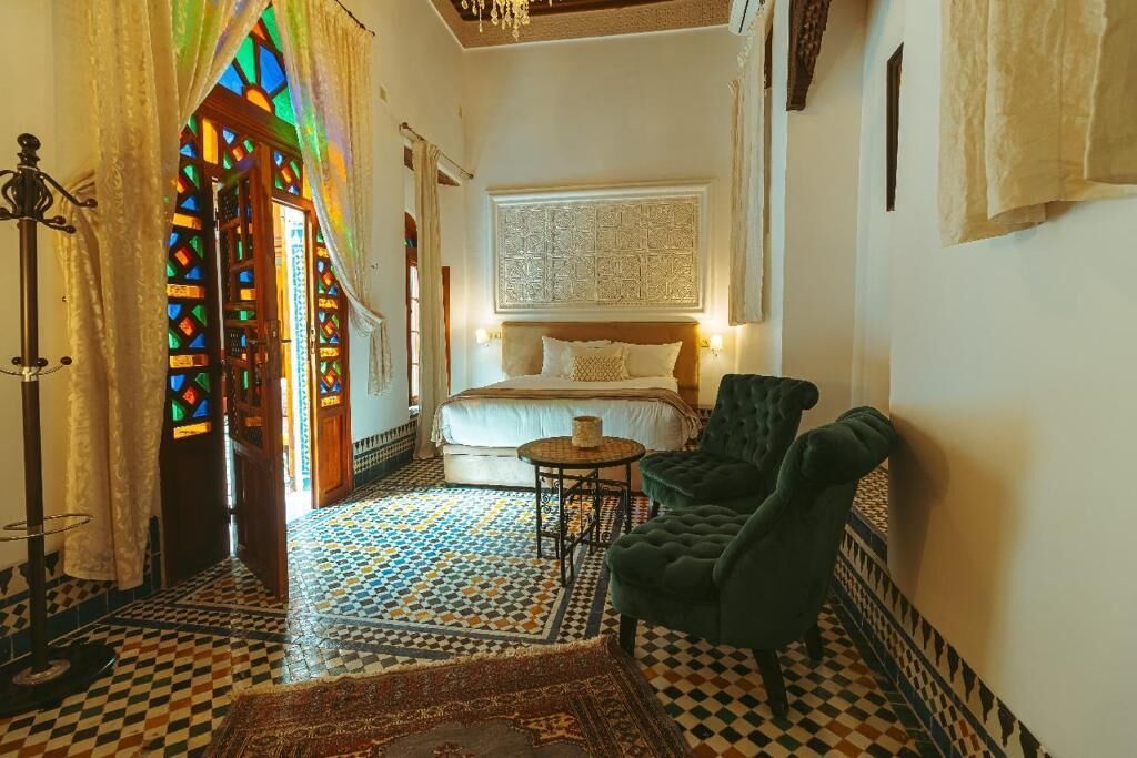 A room with a bed, a small round table, two green armchairs, a patterned floor, and a decorative wall panel.