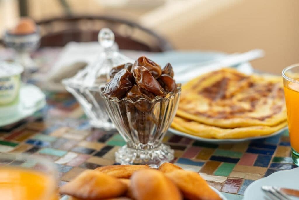 A breakfast setting with a glass bowl of chocolate, a plate of croissants, a pizza, and a glass of orange juice on a tiled table.