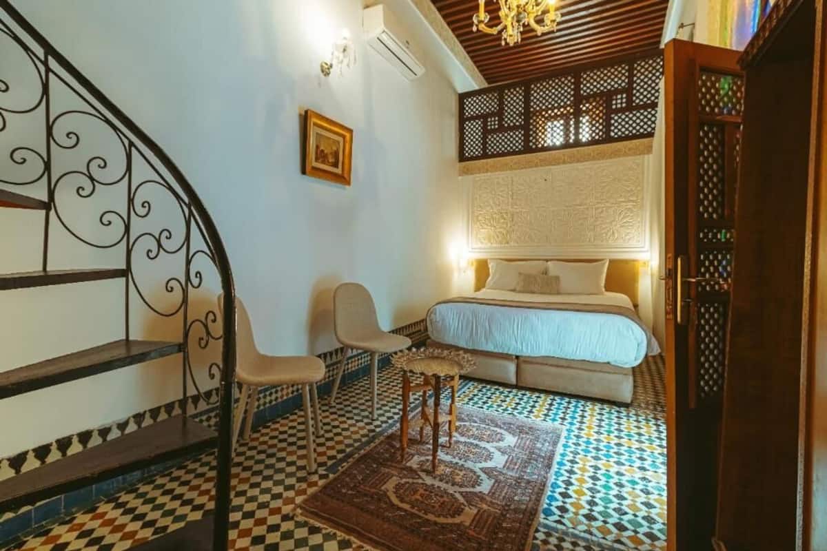 A bedroom with a bed, a chair, a small table, a patterned rug, and a decorative wall panel.