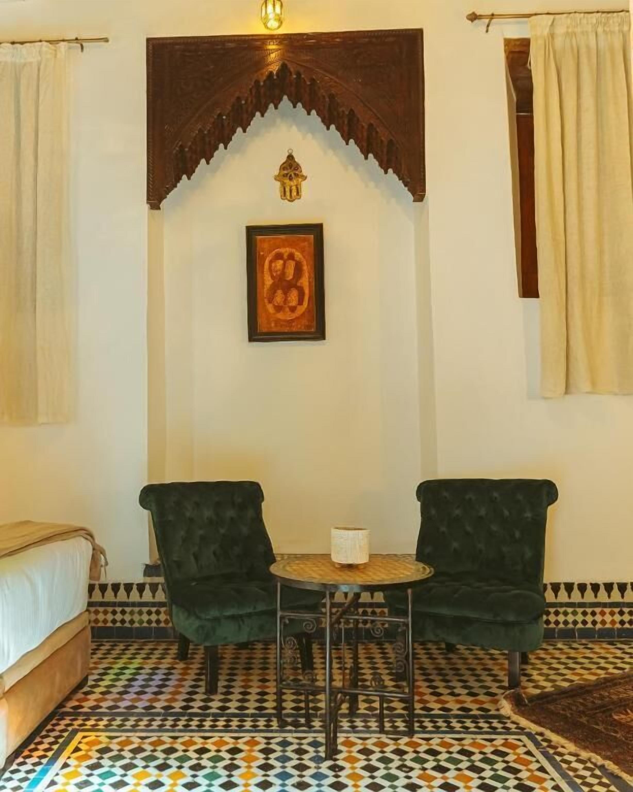 A room with a bed, two chairs, a small table, a patterned floor, a framed picture, and a decorative arch.