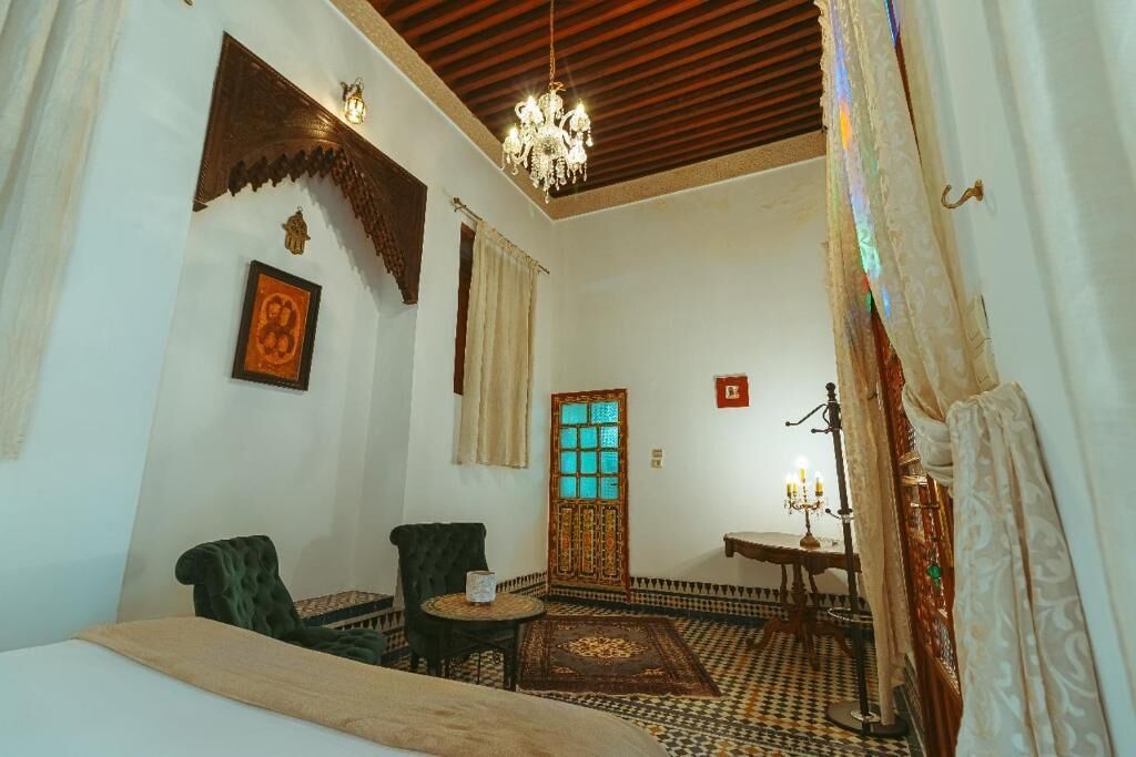 A room with a bed, two chairs, a small table, a chandelier, a patterned rug, and a door with stained glass.