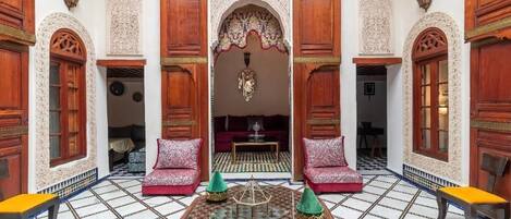 Un interior tradicional marroquí con puertas de madera ornamentadas, una zona de estar central con cojines y un suelo de mosaicos.