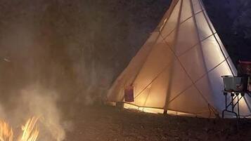 Uma tenda tipi com uma fogueira na frente, em meio a um chão de terra.