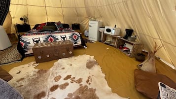 Interior de una carpa con una cama, un baúl de almacenamiento, un refrigerador blanco, un microondas y una pequeña mesa con objetos.