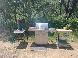 An outdoor cooking area with a portable grill, a cooler, and a small table with utensils.