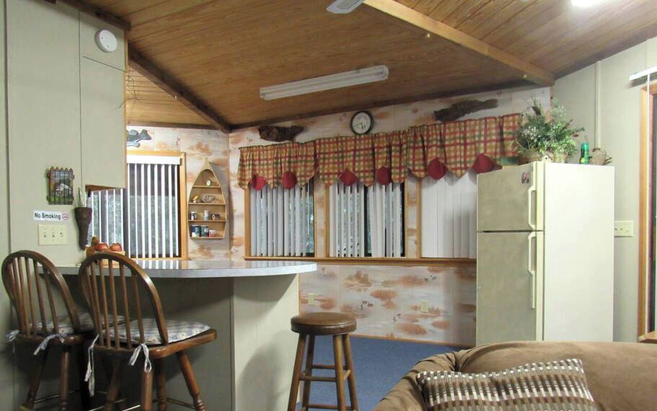 A kitchen with a wooden ceiling, a refrigerator, a dining area with two chairs, a bar counter, and a clock on the wall.