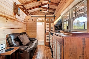 Interior de una cabaña con detalles de madera, un sofá de cuero, una mesita y una ventana con vistas al exterior.