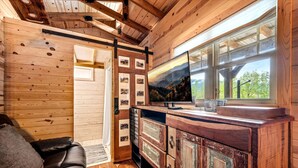 A cabin interior with a wooden ceiling, a TV on a wooden cabinet, a leather sofa, and a window with a view of trees. - Appalachian Adventure in this Tiny Cabin near Hiawassee, Ideal for Couples' Glamping (Hayesville)